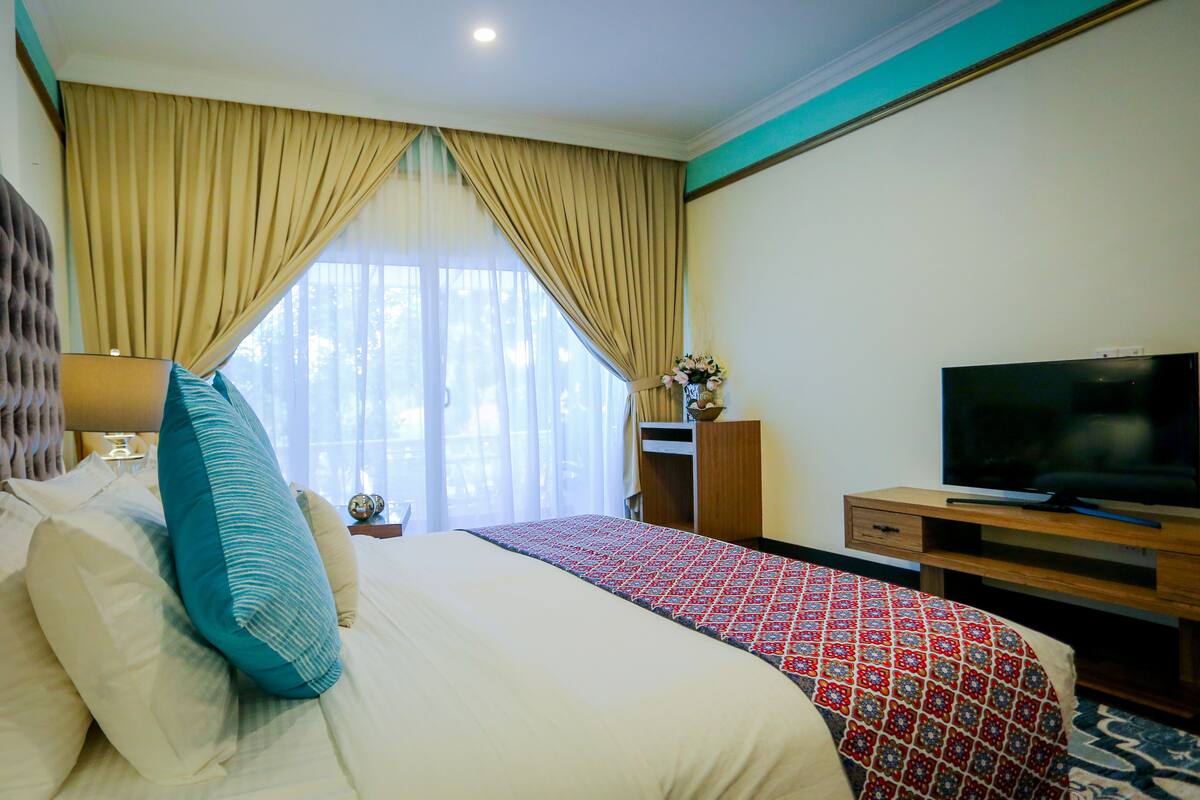A hotel room with a large bed, a TV, a desk, and a window with curtains.