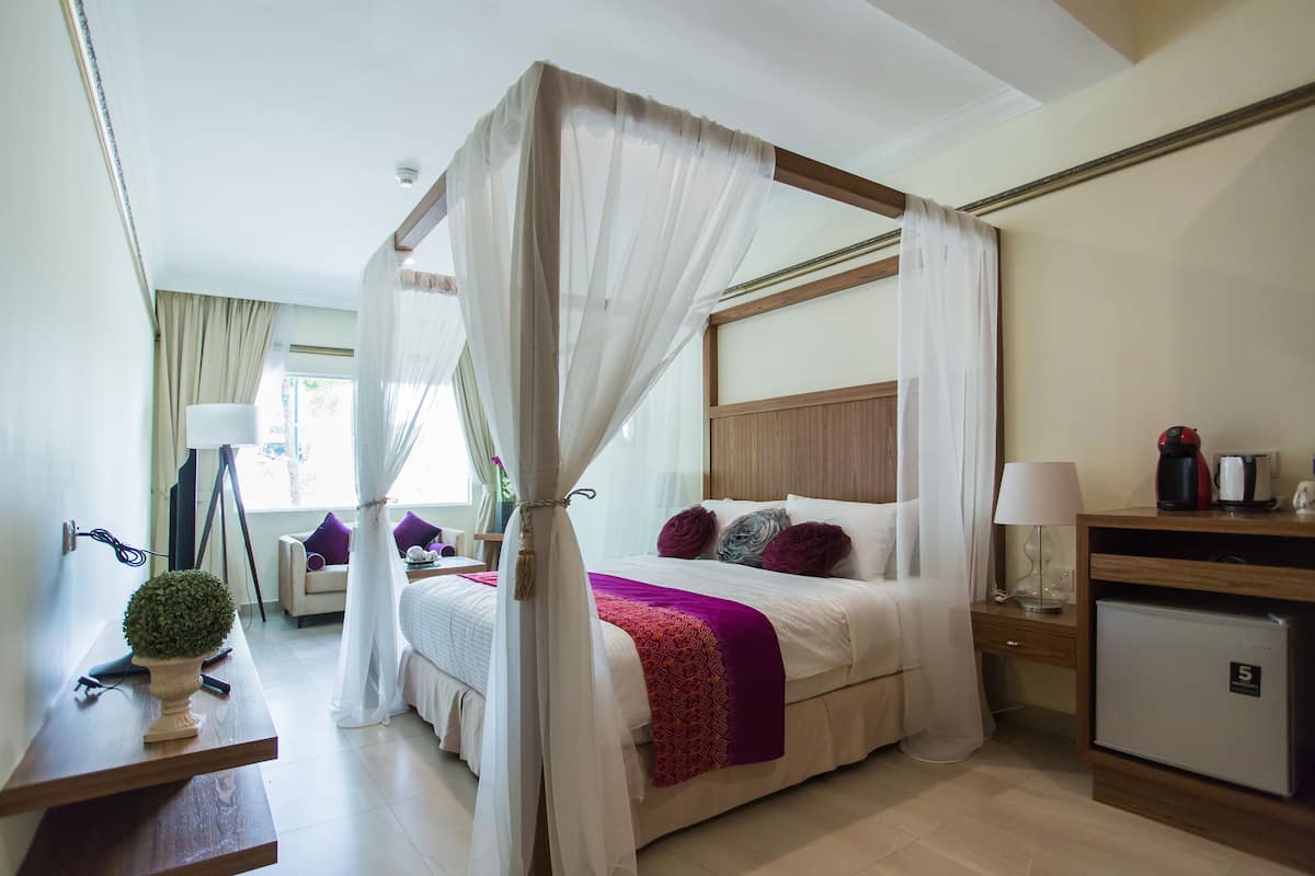 A four-poster bed with a canopy, a bedside table with a lamp, a small refrigerator, and a desk with a plant.