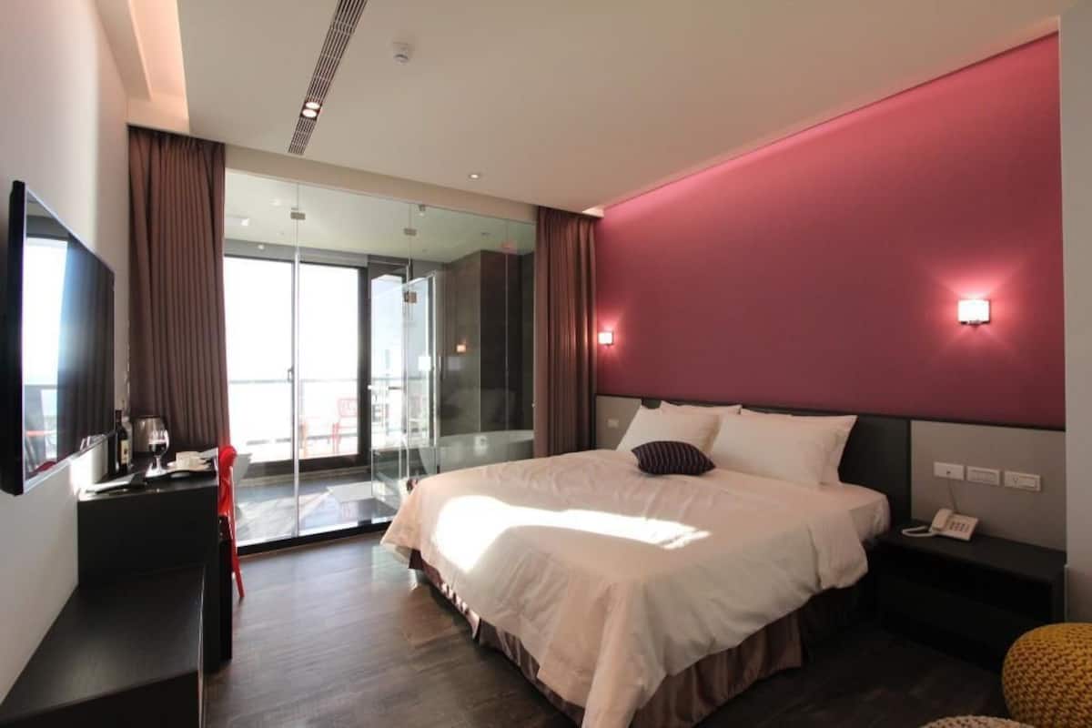A modern hotel room with a large bed, a desk, a chair, a glass door leading to a balcony, and a red accent wall.