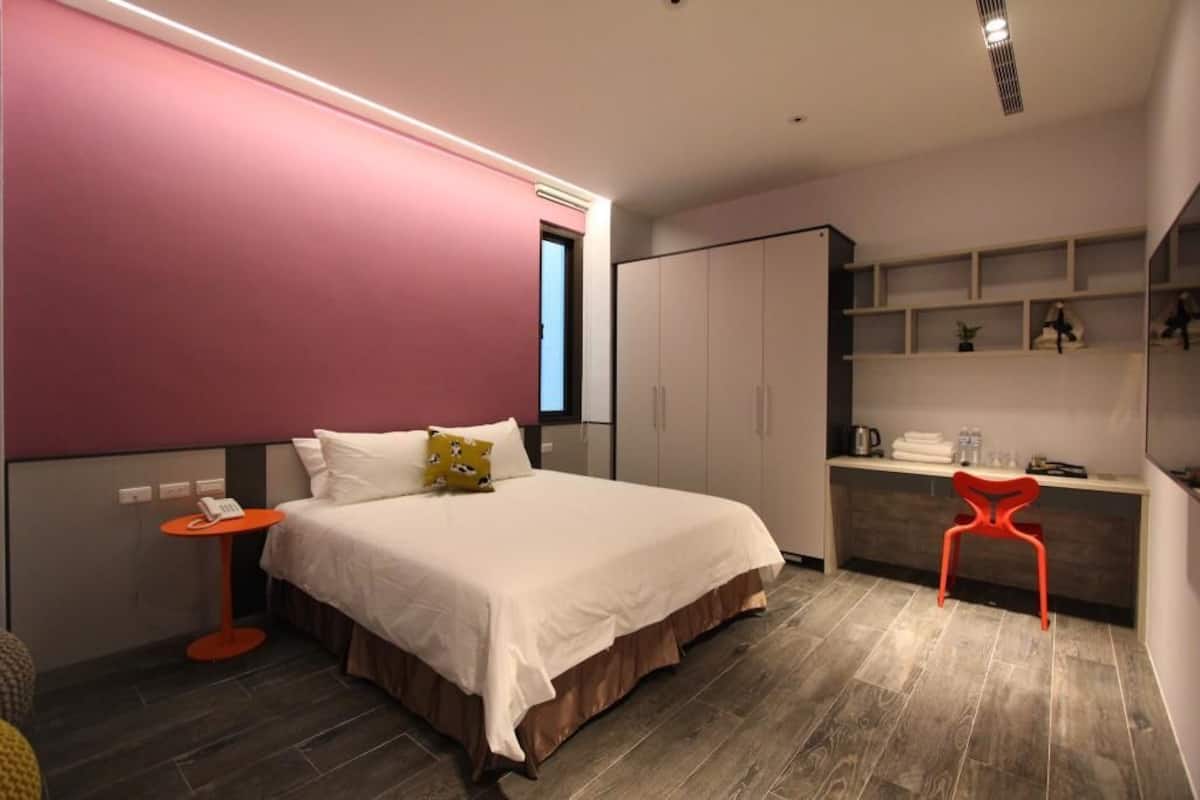 A modern hotel room with a large bed, a small table, a red chair, and built-in shelving with toiletries.
