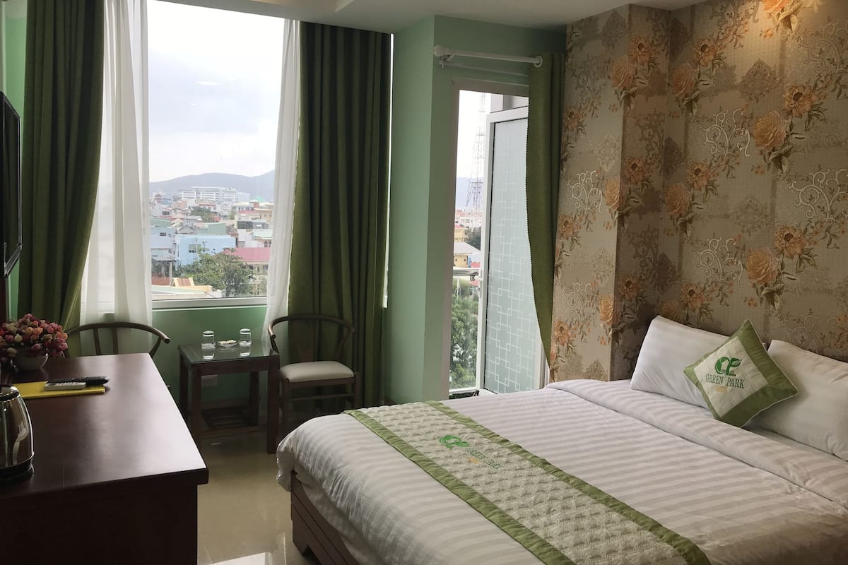Ein Hotelzimmer mit Bett, Schreibtisch, Stuhl und Blick auf die Stadt durch das Fenster sowie einem Balkon.