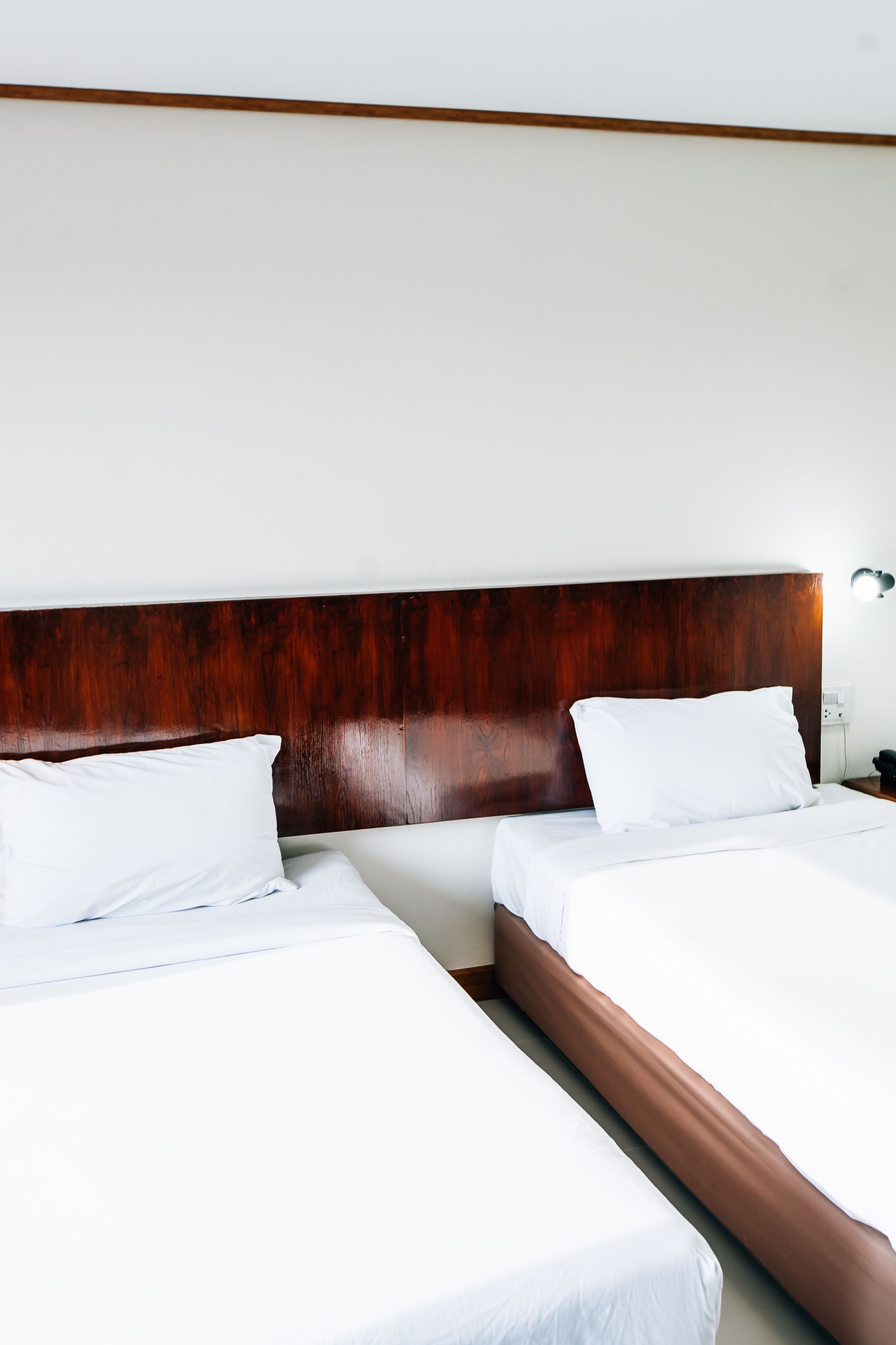 A hotel room with two single beds, wooden headboards, white linens, and a bedside table with a lamp.