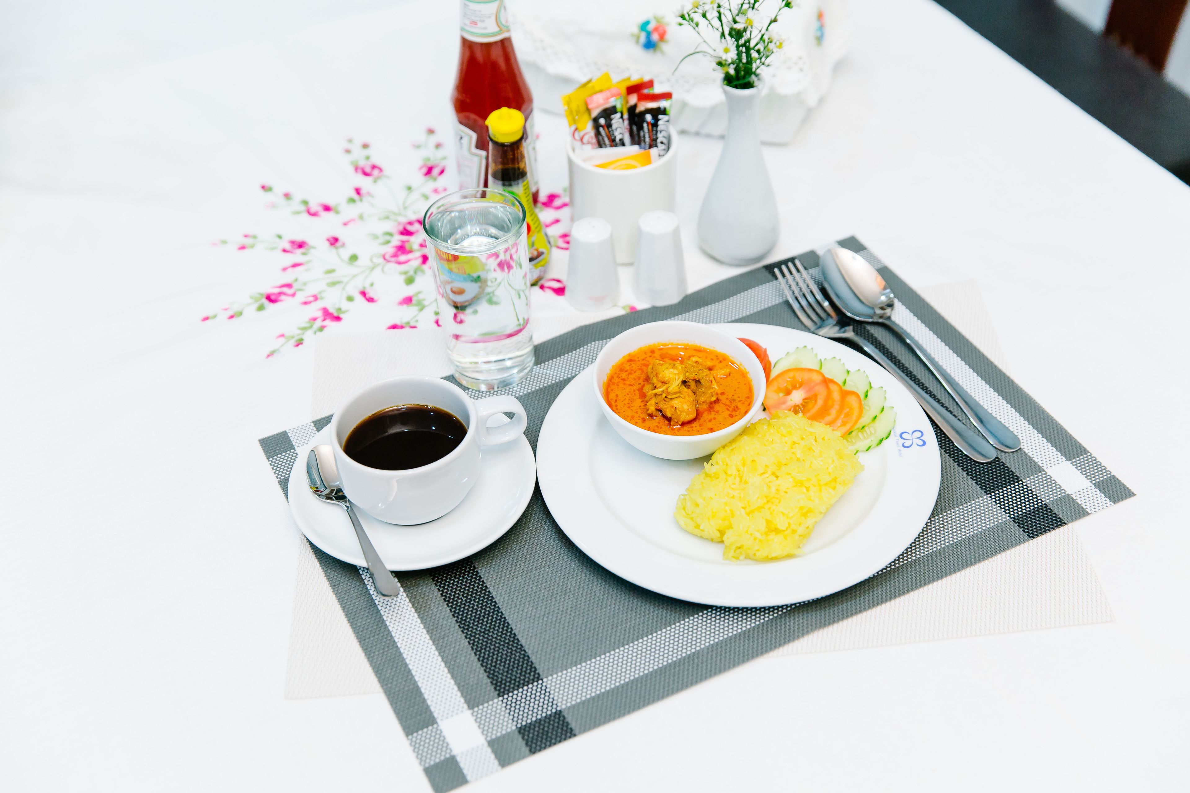 Una mesa dispuesta con comida que incluye arroz, verduras y un tazón de sopa, además de una taza de café y salsas.