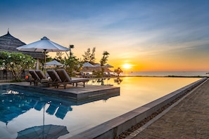 Un espace aménagé au bord de la piscine, avec des chaises longues et des parasols, offrant une vue sur une étendue d’eau calme au coucher du soleil.