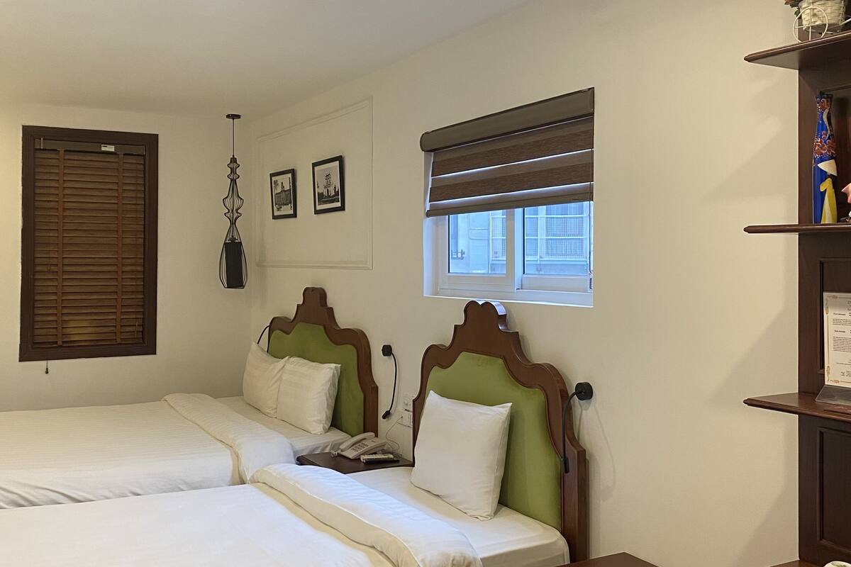 A hotel room with two beds, a small table, a nightstand, a window with blinds, a wall-mounted phone, and a shelf with decorative items.