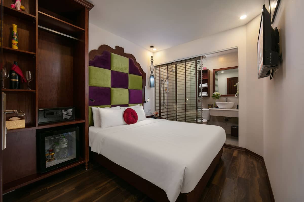 A hotel room with a large bed, a wardrobe, a minibar, a television, and a bathroom with a sink and mirror.