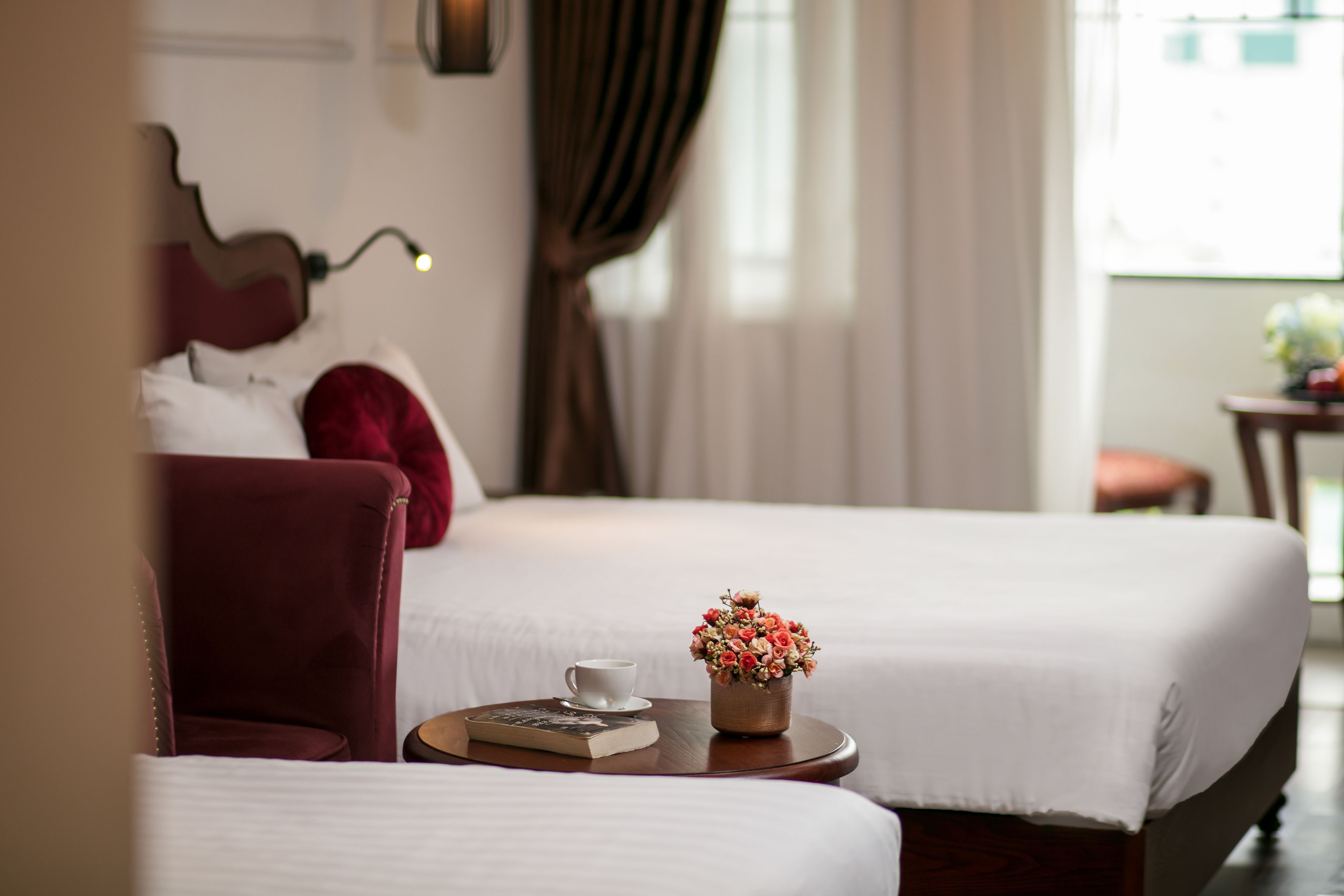 A hotel room with two beds, a small table with a cup and a book, and a vase with flowers.
