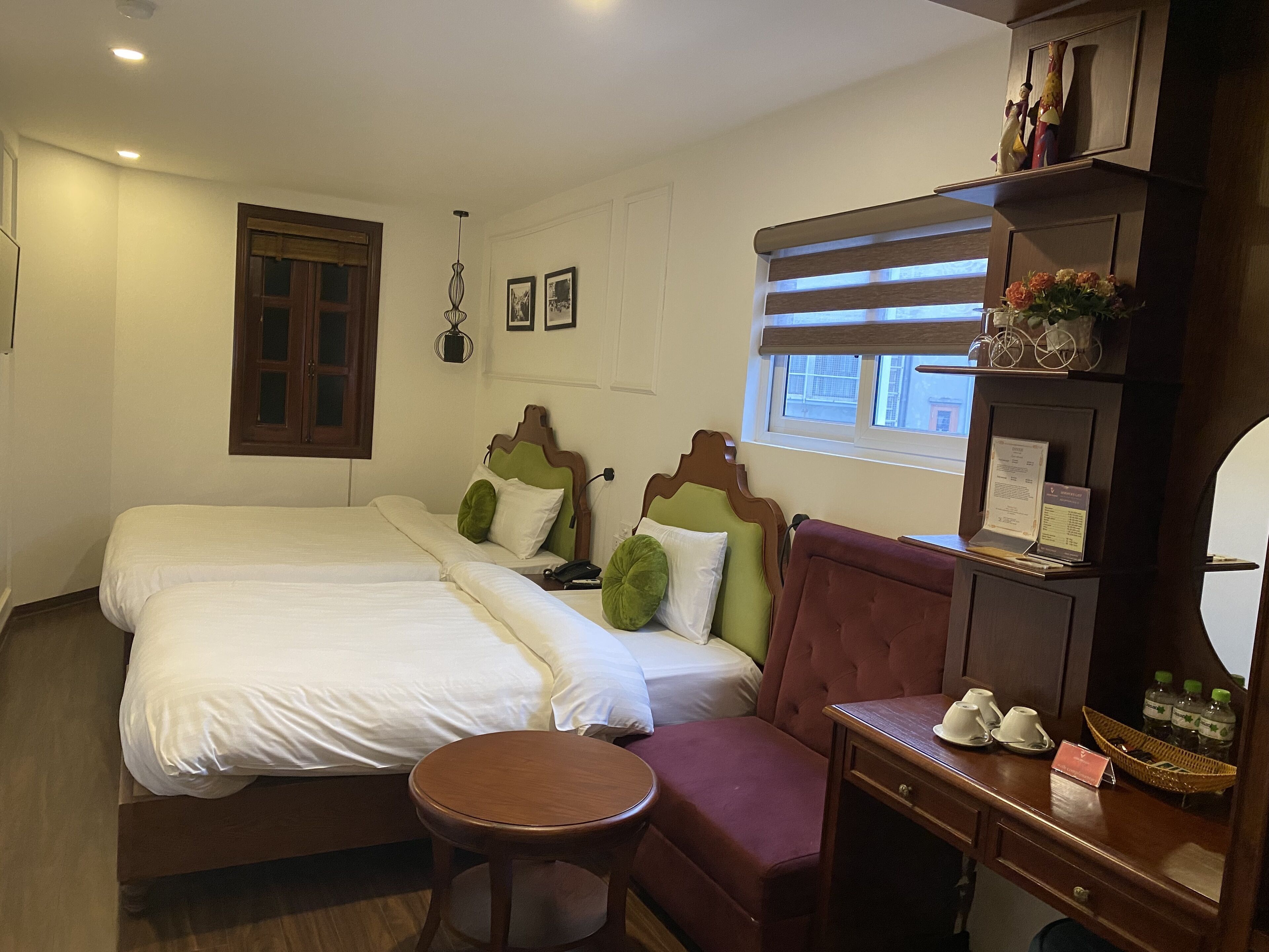 A hotel room with two beds, a small table, a desk with a basket of toiletries, and a window with blinds.
