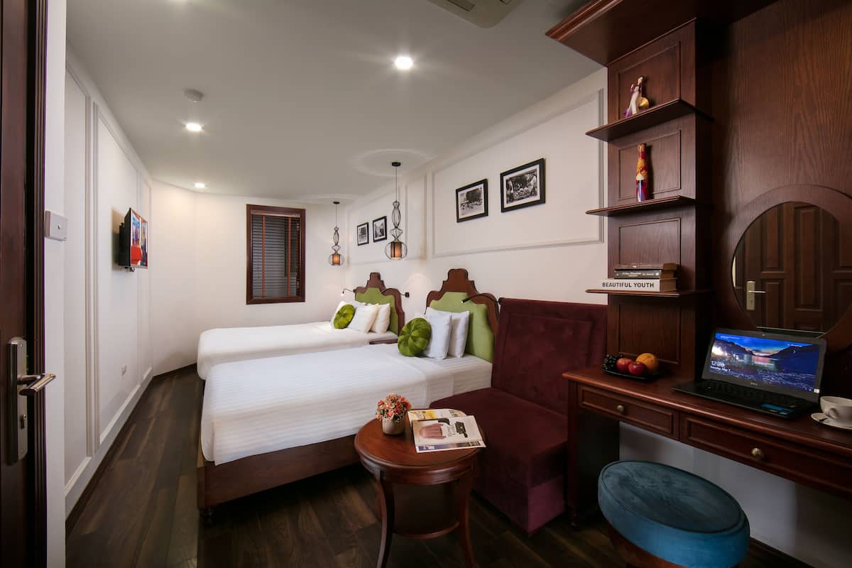 A hotel room with two beds, a desk with a laptop, a small table with a vase and a magazine, and a blue chair.