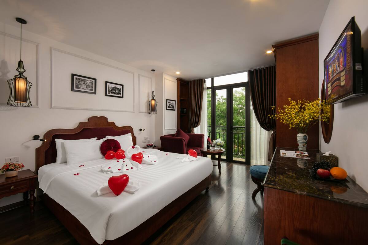 A hotel room with a large bed, a desk, a chair, a window with curtains, and a vase with flowers.