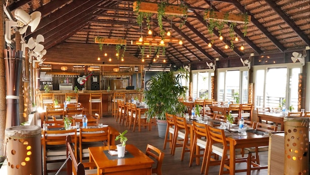Un restaurante espacioso con mesas y sillas de madera, plantas colgantes e iluminación colgante.