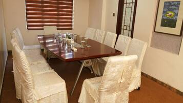 A conference room with a long table, white chairs with lace covers, a window with blinds, and a framed picture on the wall.