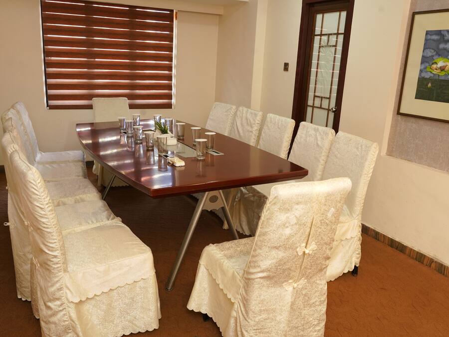 A conference room with a long table, white chairs with lace covers, a window with blinds, and a framed picture on the wall.