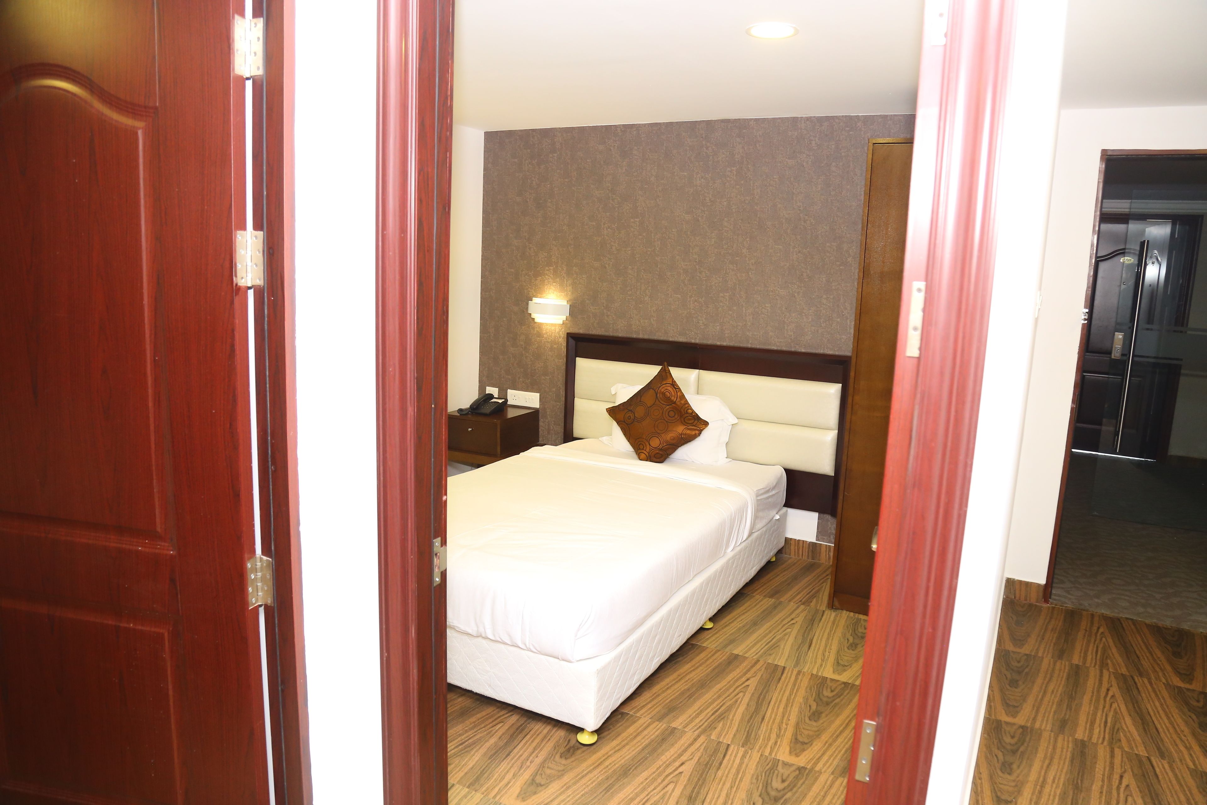 Una habitación de hotel con piso de madera, una cama blanca, una mesita de noche y una lámpara de pared.