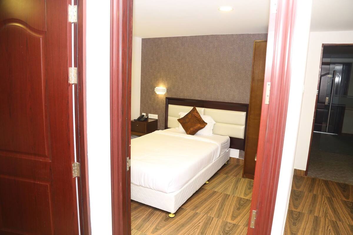 Een hotelkamer met een bed, nachtkastje, lamp en een deur naar een andere kamer.