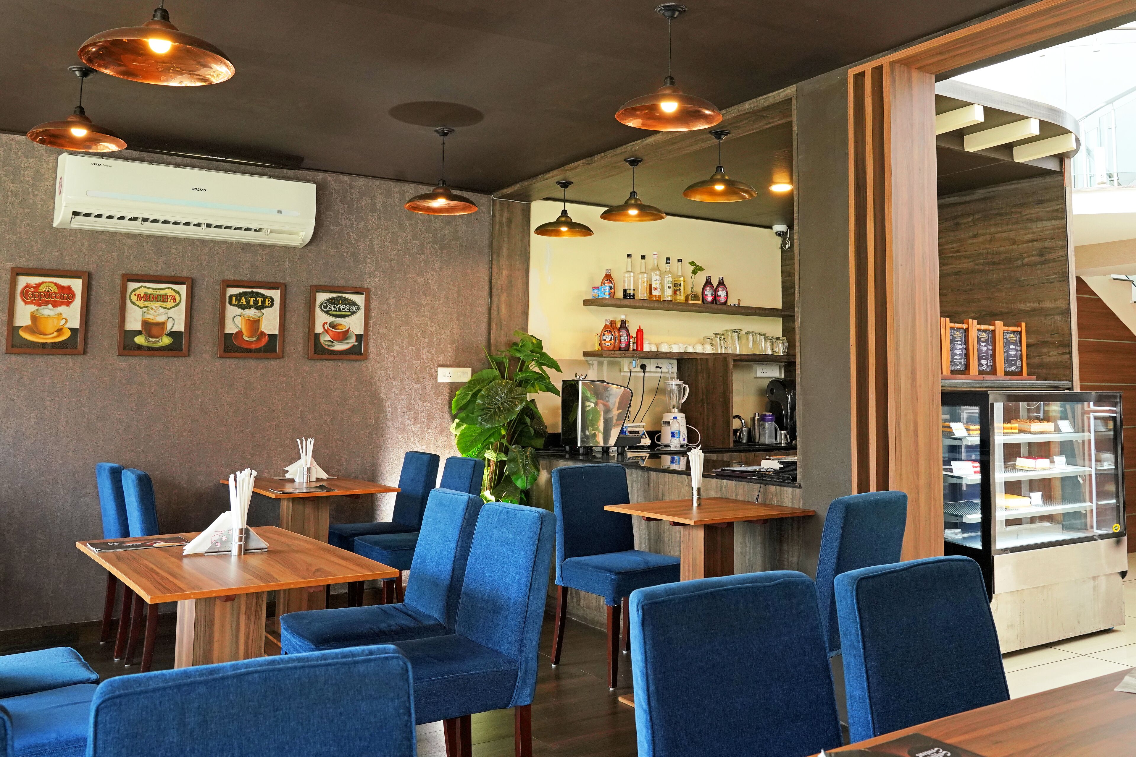 A restaurant interior with blue upholstered chairs, wooden tables, hanging copper pendant lights, and a display case with desserts.