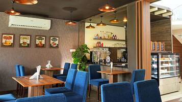 A restaurant interior with blue upholstered chairs, wooden tables, hanging copper pendant lights, and a display case with desserts.