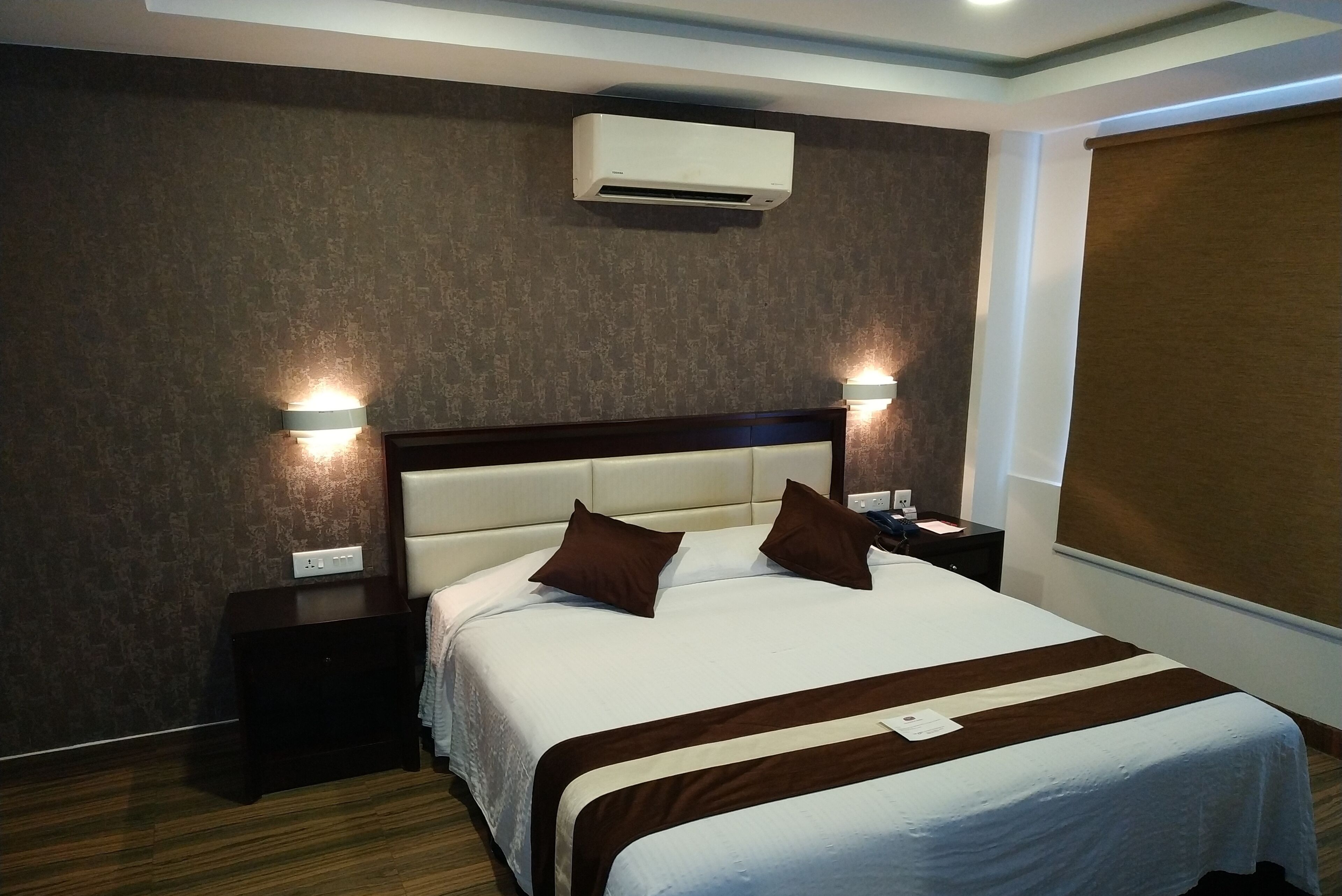 A hotel room with a large bed, two bedside tables, an air conditioning unit, and a wall-mounted light fixture.