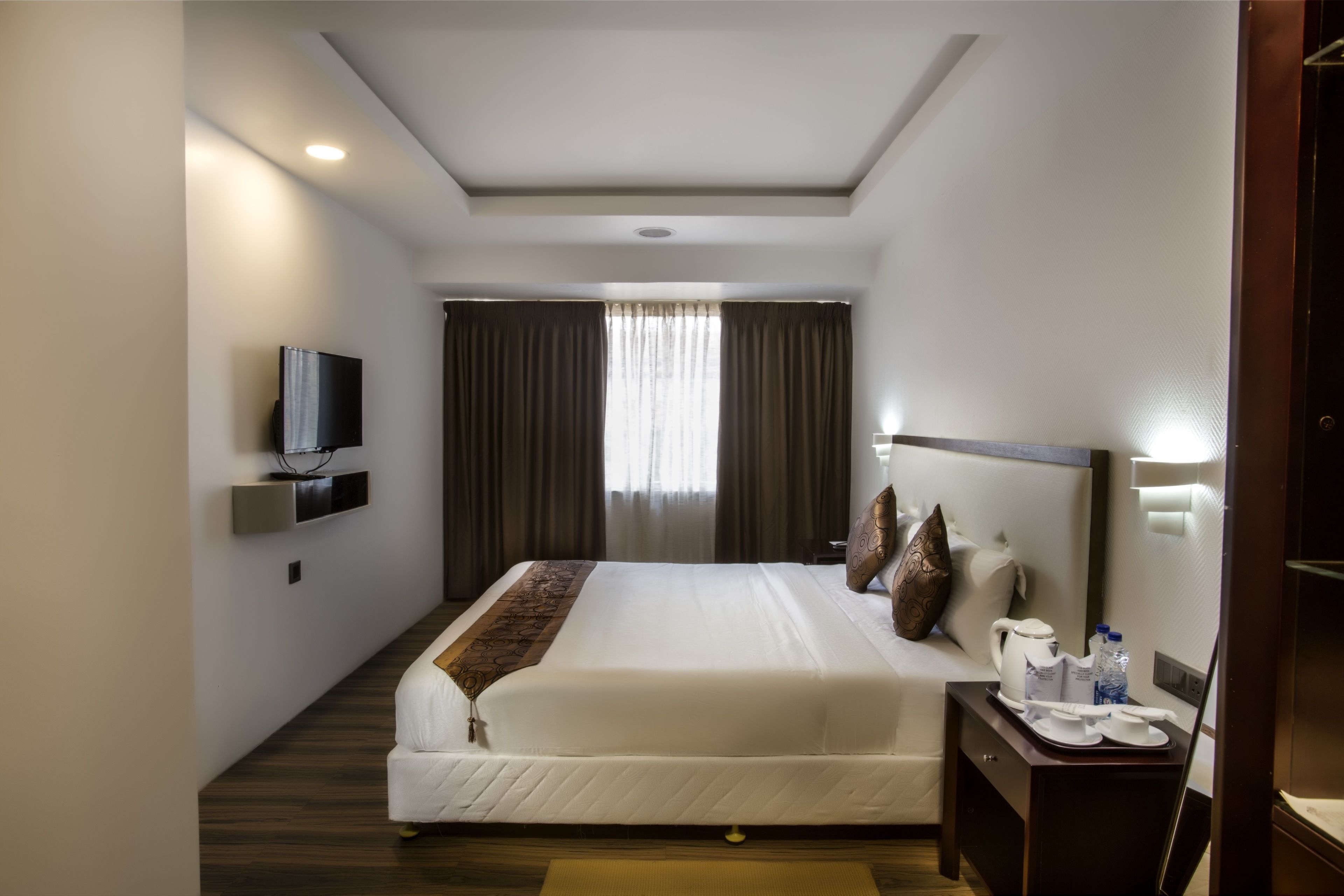A hotel room with a large bed, a flat-screen TV mounted on the wall, a bedside table with a tray of breakfast items, and a window with curtains.