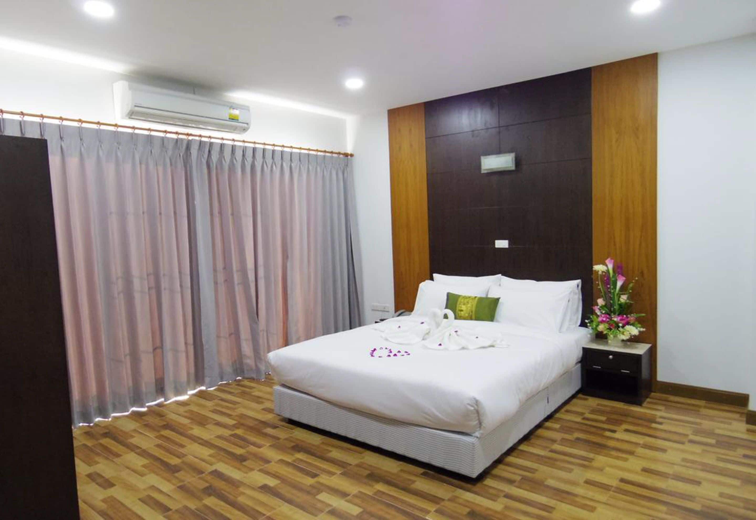 A hotel room with a large bed, wooden flooring, a nightstand with a vase of flowers, and a wall-mounted air conditioner.