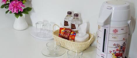 Una mesa con un jarrón blanco lleno de flores rosadas, una cesta con agua embotellada y aperitivos, un hervidor eléctrico blanco y dos vasos vacíos.