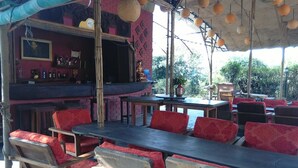 An outdoor seating area with wooden tables and red cushioned chairs, a bar area with bottles and a Buddha statue, and a view of greenery in the background. - Lake Twenty Thousand Farm and Retreat (Bharatpur)
