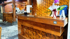 Um balcão de atendimento com design de madeira, uma placa com a inscrição 'Front Desk', uma orquídea roxa decorativa e uma placa indicando 'Não aceitamos dinheiro'.