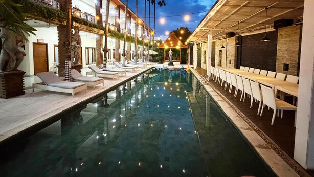 Uma área junto à piscina com espreguiçadeiras, um espaço de refeições e um edifício com colunas e varanda.