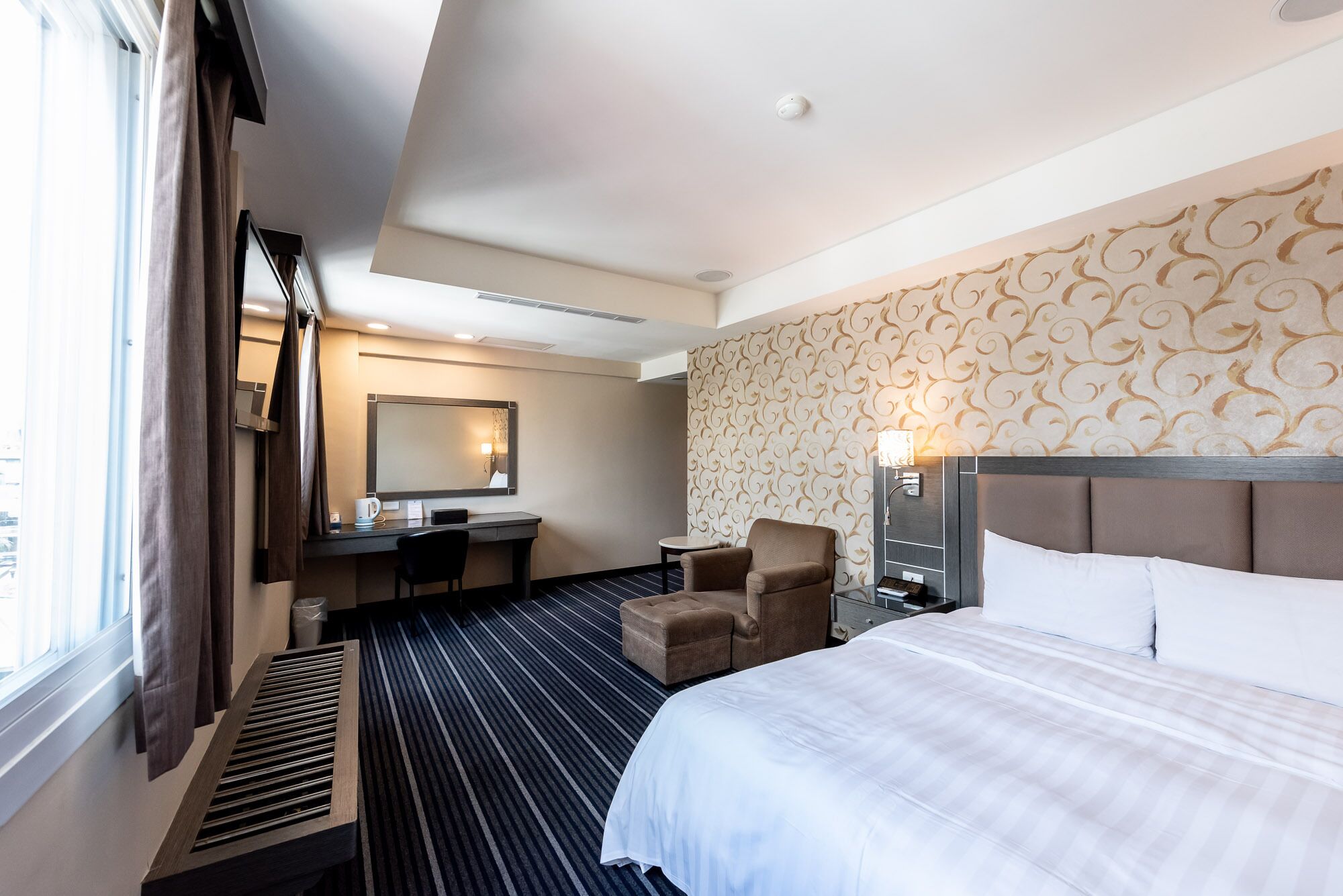 A hotel room with a bed, a desk, a chair, a small table, and a large mirror.