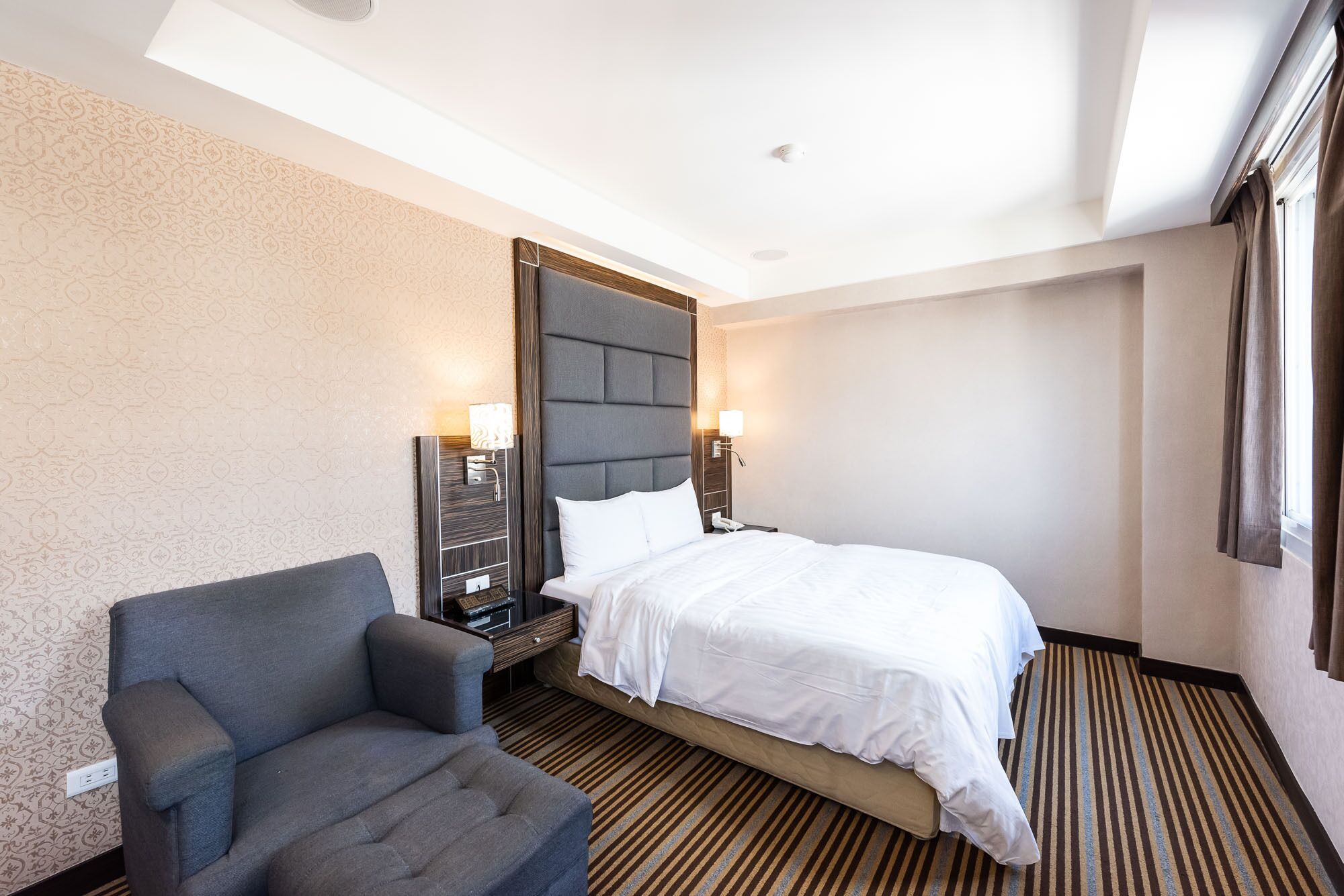 A hotel room with a bed, a chair, a nightstand, and a wall-mounted lamp.