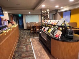 Een hotel receptie met een balie waarop boeken en een koffiemachine staan. Er is een zithoek met tafels en stoelen, en een projectiescherm op de achtergrond.