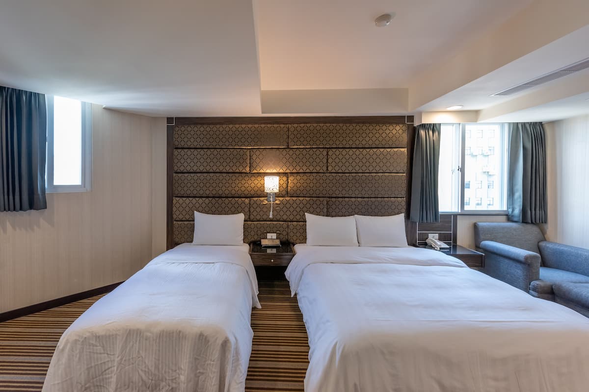 A hotel room with two beds, a patterned headboard, a bedside table with a lamp, and a view of the city through the window.