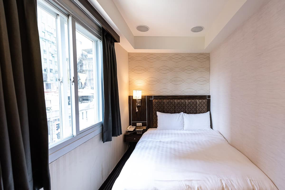 A hotel room with a large window, a bed with white linens, a nightstand with a lamp, and a patterned wall.