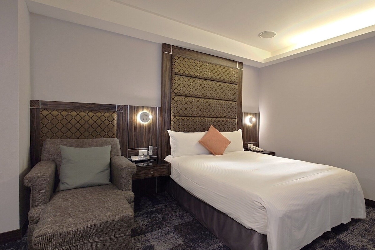 A hotel room with a large bed, a chair, a nightstand, and a wall-mounted light fixture.