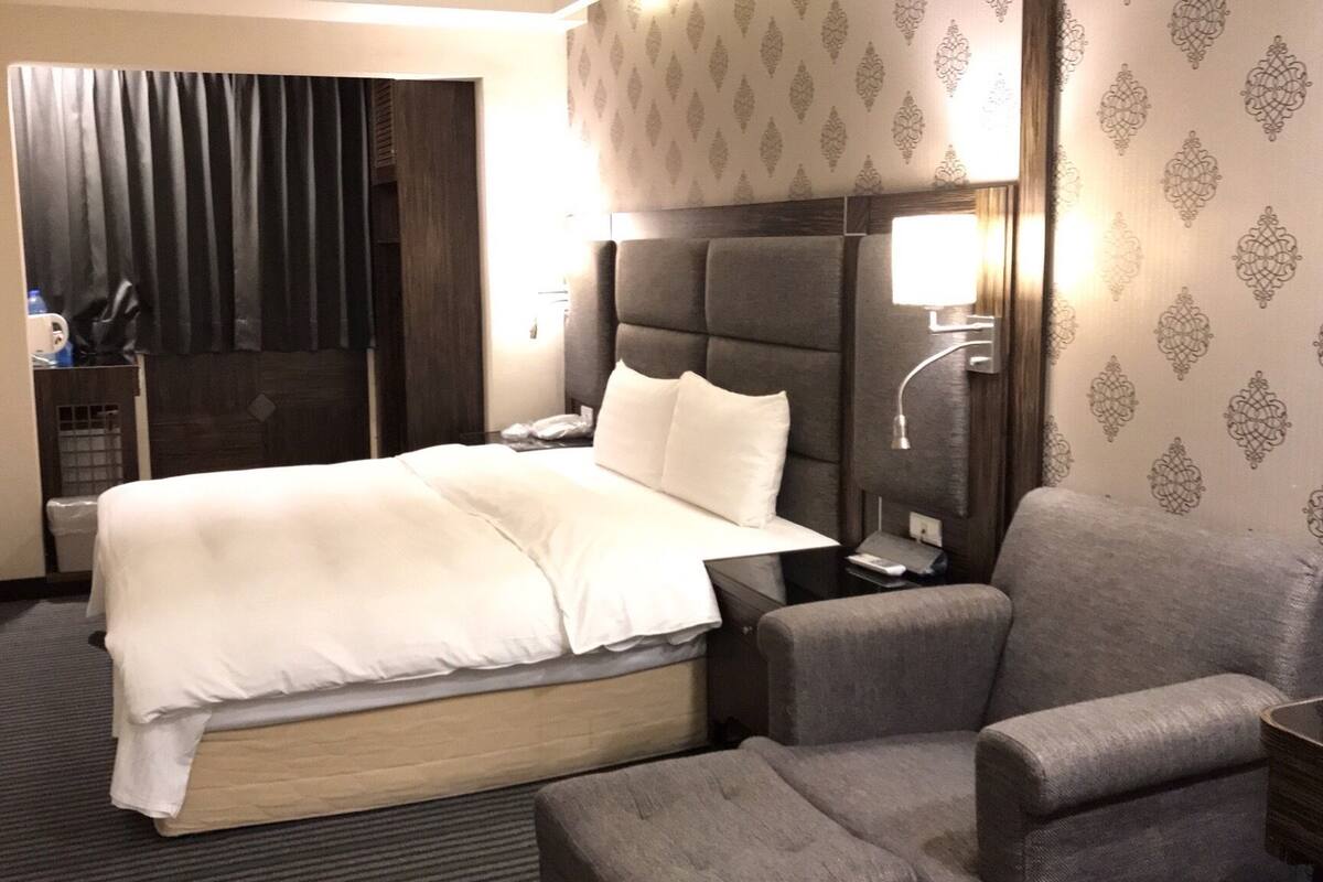 A hotel room with a large bed, a grey armchair, a bedside table with a lamp, and a TV mounted on the wall.