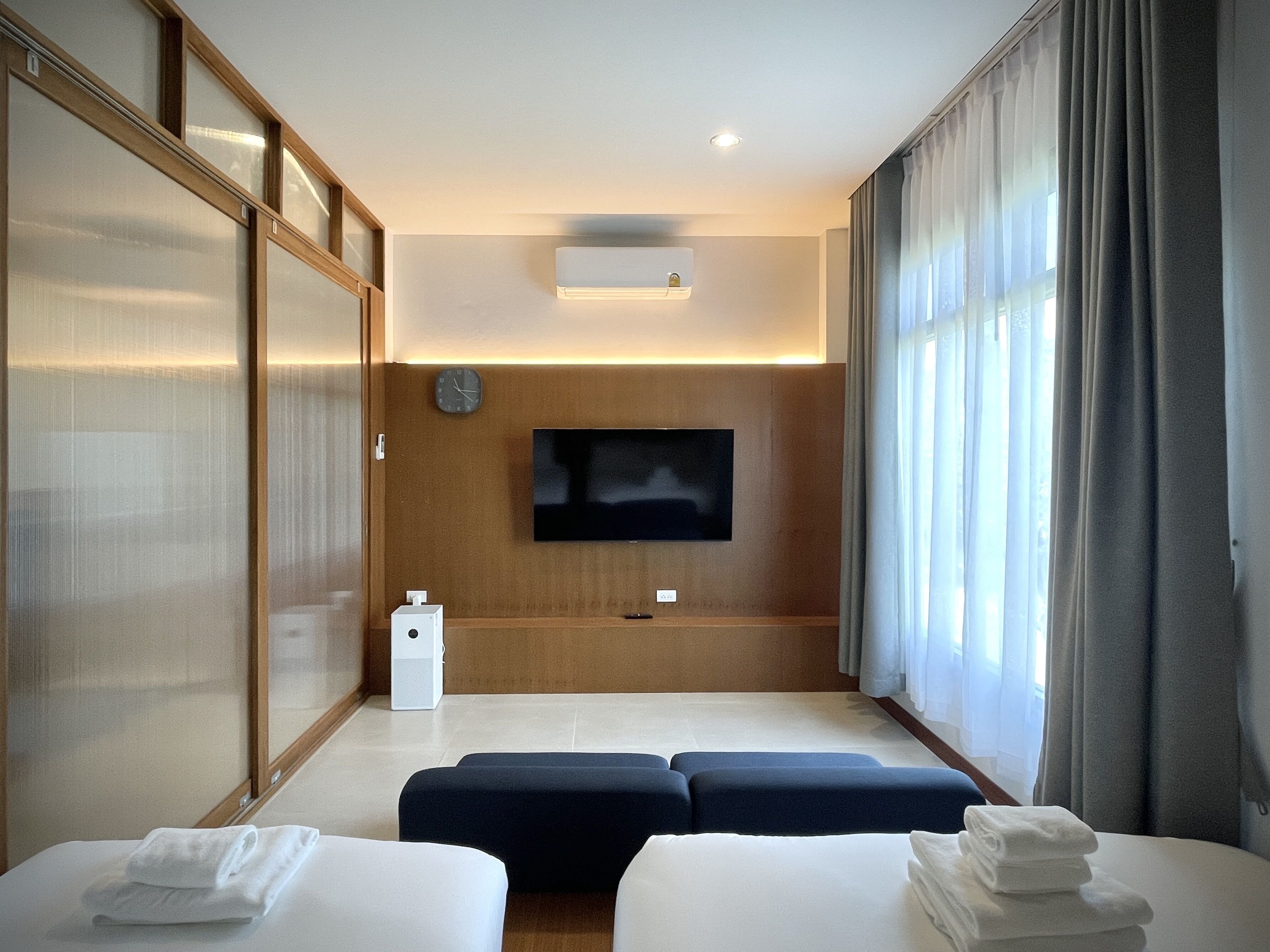 A modern hotel room with a flat-screen TV mounted on the wall, a wooden panel wall, a large window with curtains, and two beds with towels.