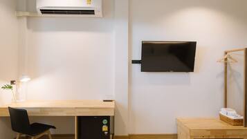 A modern room with a wooden desk, a black chair, a TV mounted on the wall, a small heater under the desk, and a wooden cabinet with towels.