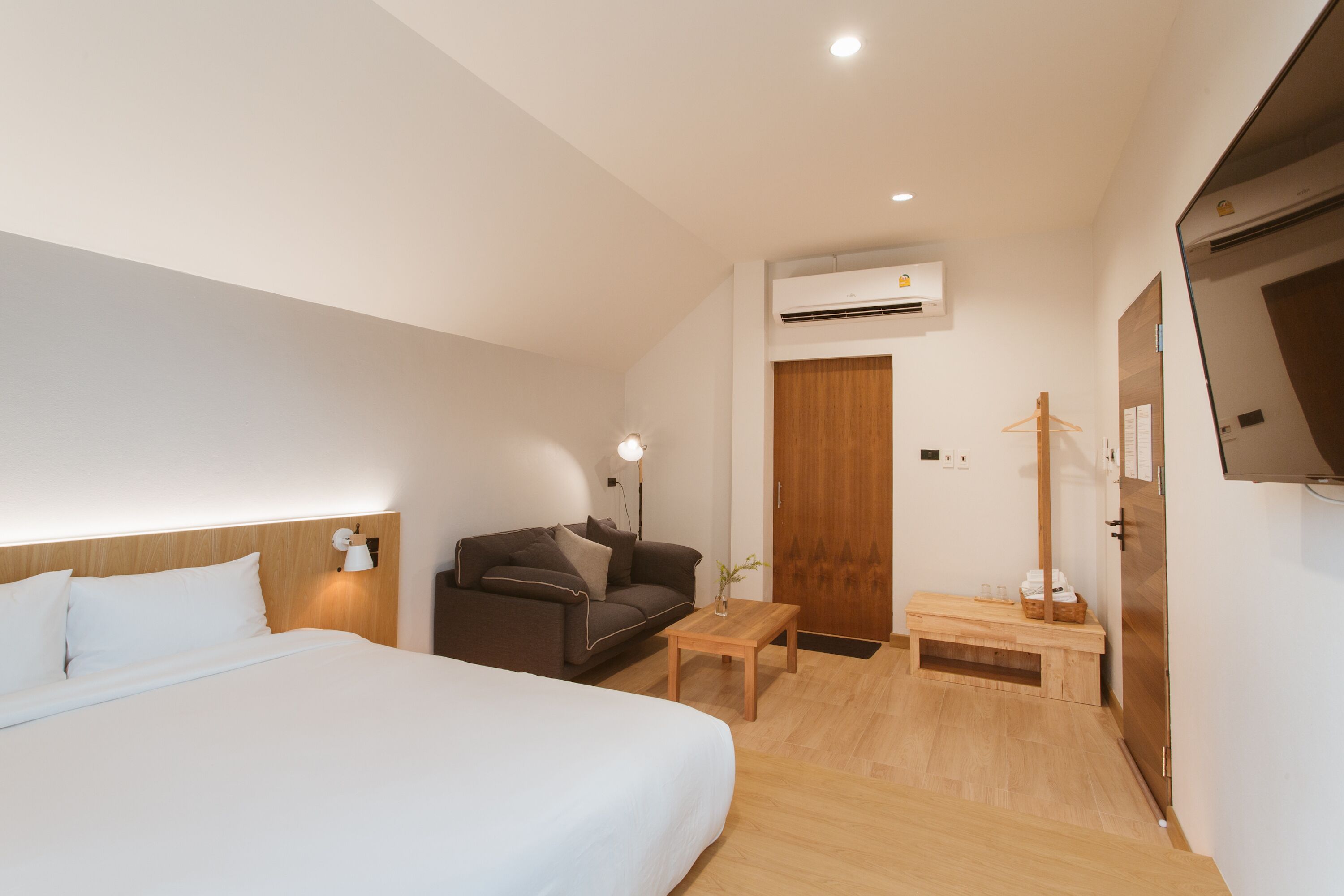 A hotel room with a bed, a sofa, a small table, a wall-mounted air conditioner, and a wooden door.