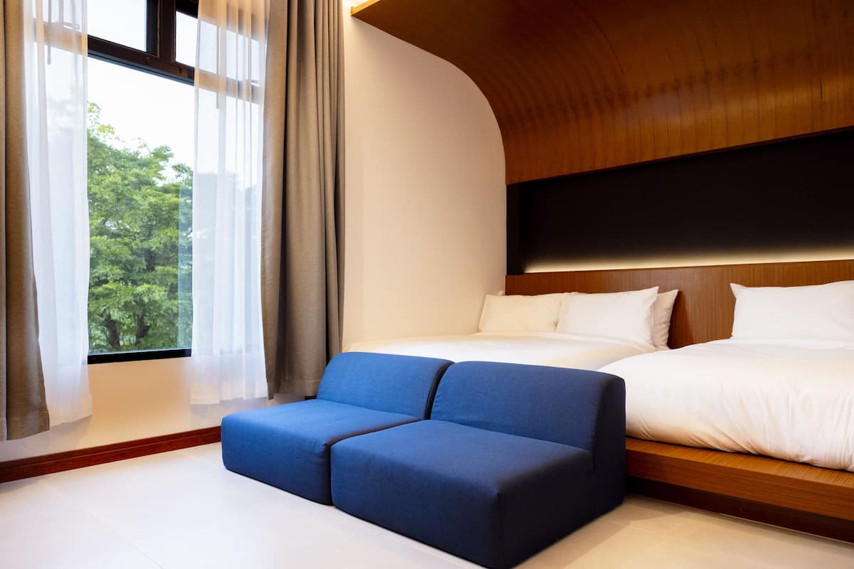 A modern hotel room with a curved wooden headboard, a large window with sheer curtains, a blue sofa, and a bed with white linens.