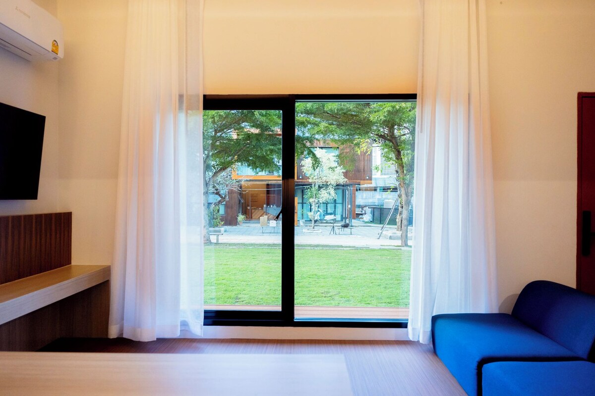 A hotel room with a bed, a blue sofa, a TV mounted on the wall, and a sliding glass door leading to an outdoor area with a lawn and trees.