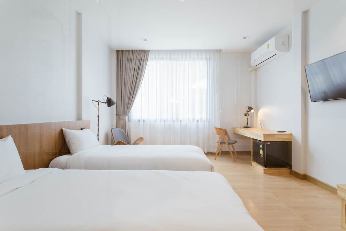 A hotel room with two beds, a desk, a chair, a TV, and an air conditioning unit.
