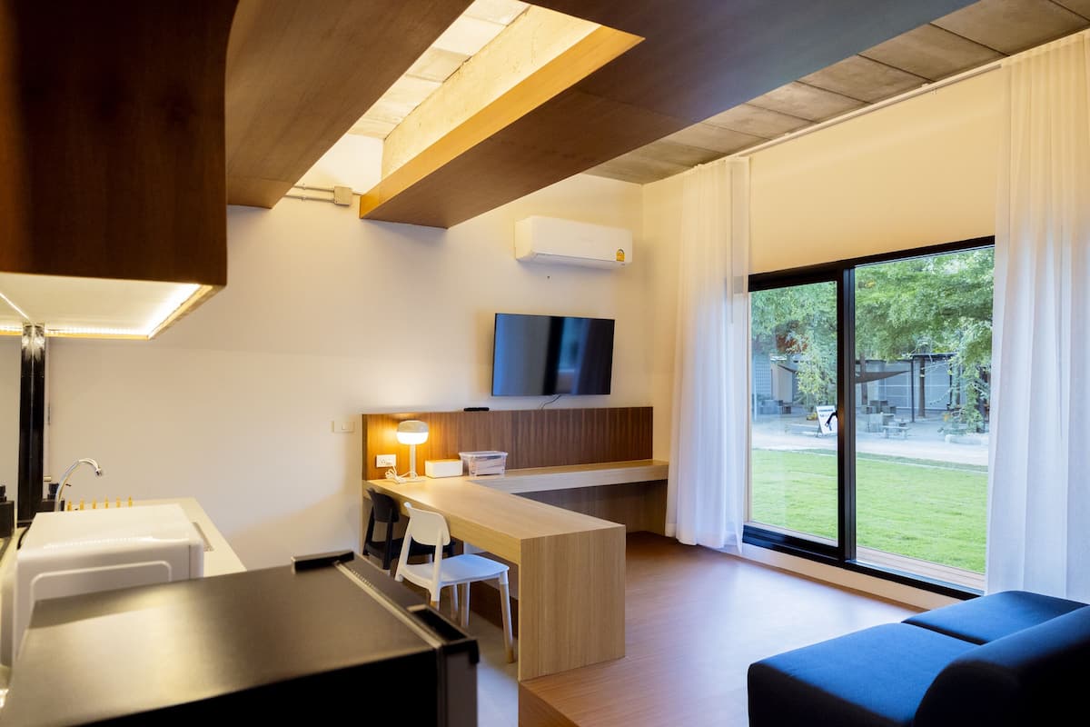 A modern room with a wooden desk, a white chair, a sofa, a TV mounted on the wall, and a large window with a view of the outdoors.