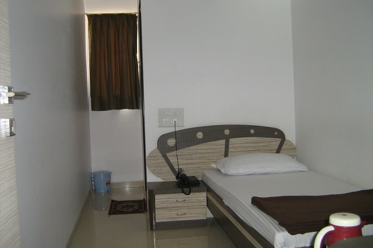 A small, simple room with a single bed, a bedside table, a nightstand, a power outlet, and a window with curtains.