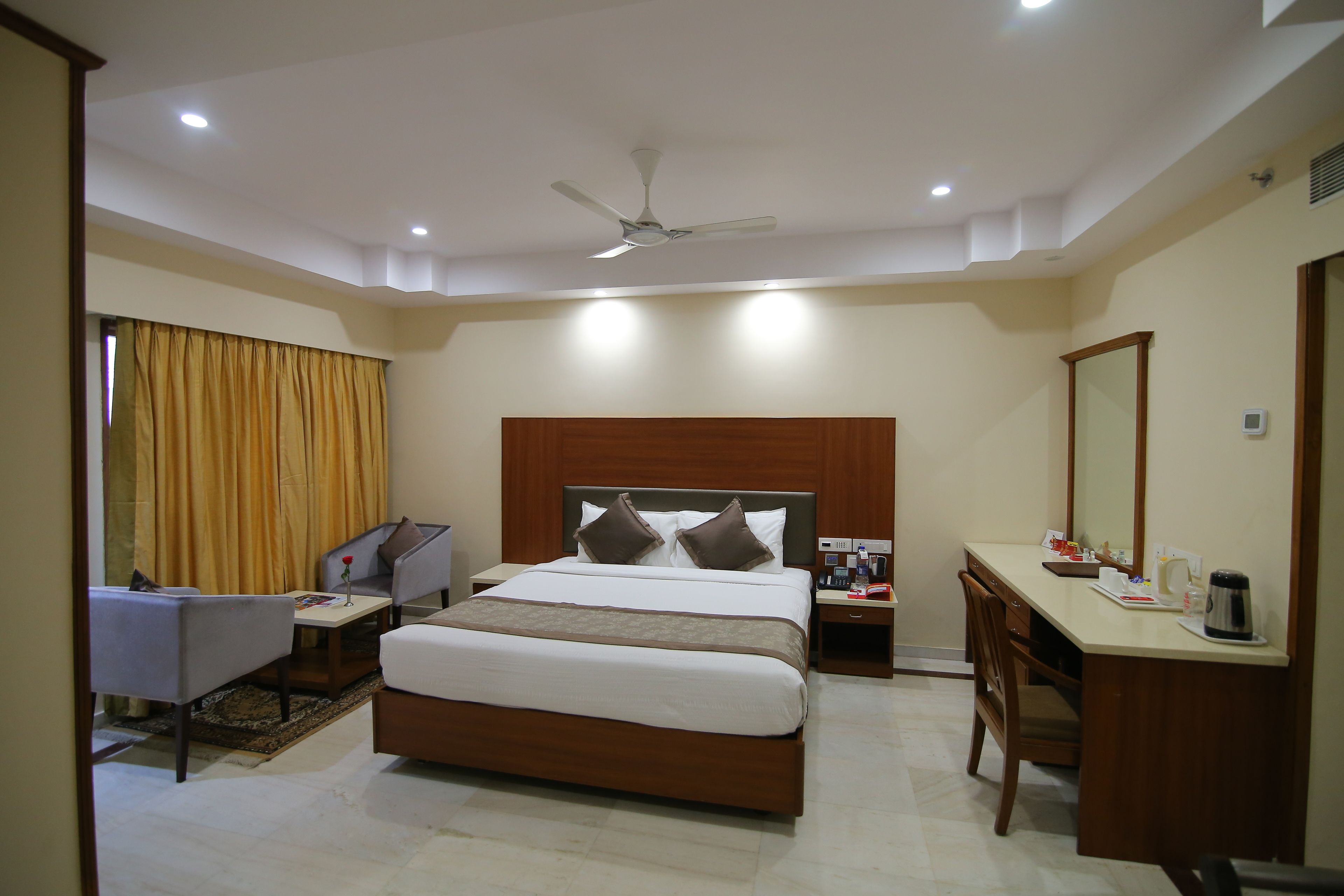 A hotel room with a bed, a desk, a chair, a nightstand, a ceiling fan, and a window with curtains.