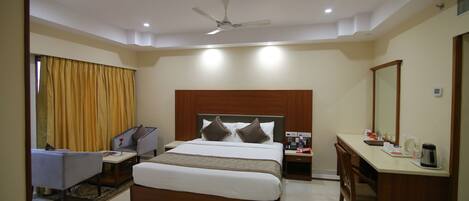 A hotel room with a bed, a desk, a chair, a nightstand, a ceiling fan, and a window with curtains.