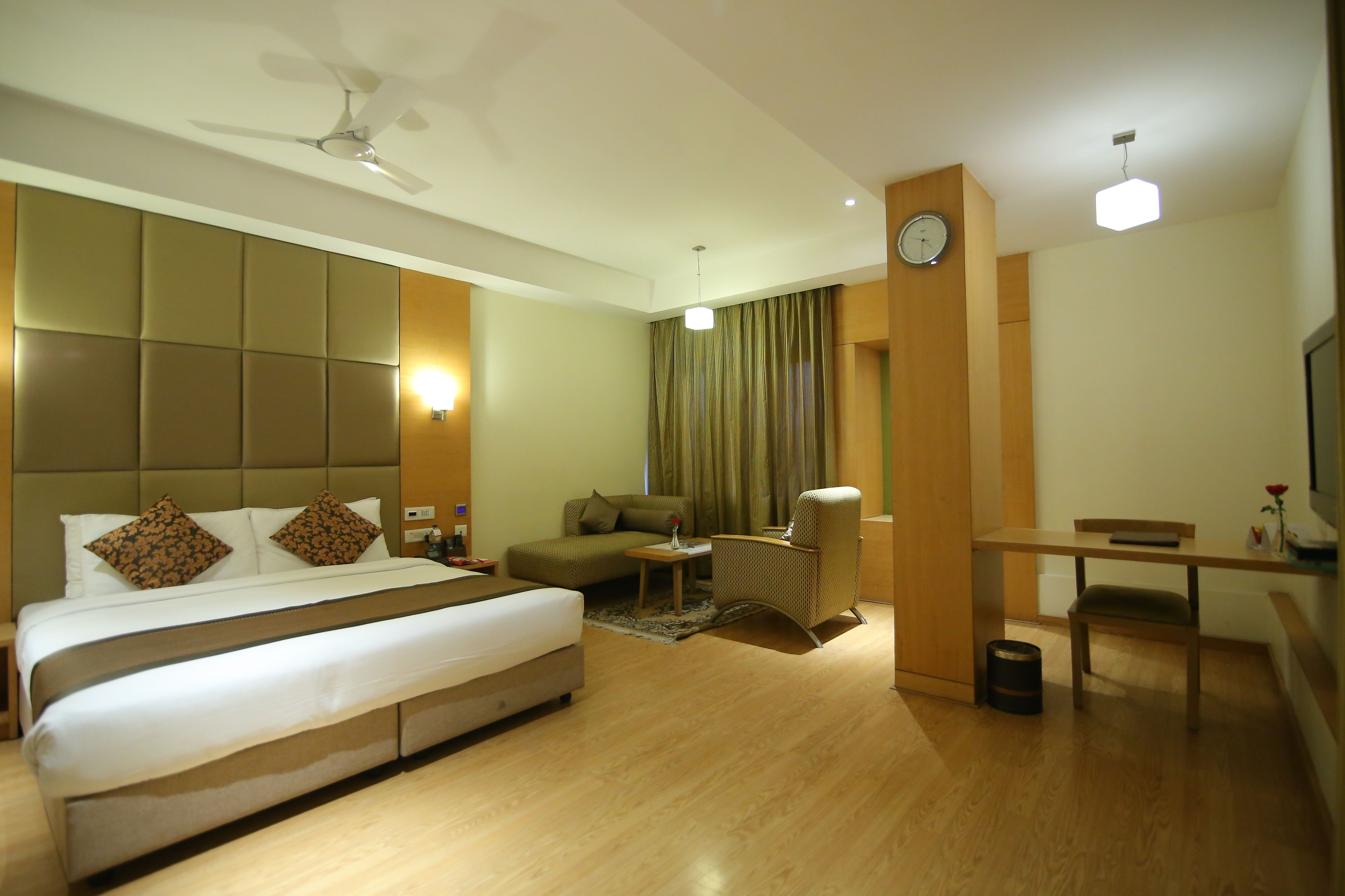 A hotel room with a large bed, a sofa, a desk, and a chair. There is a ceiling fan, a clock, and a lamp.