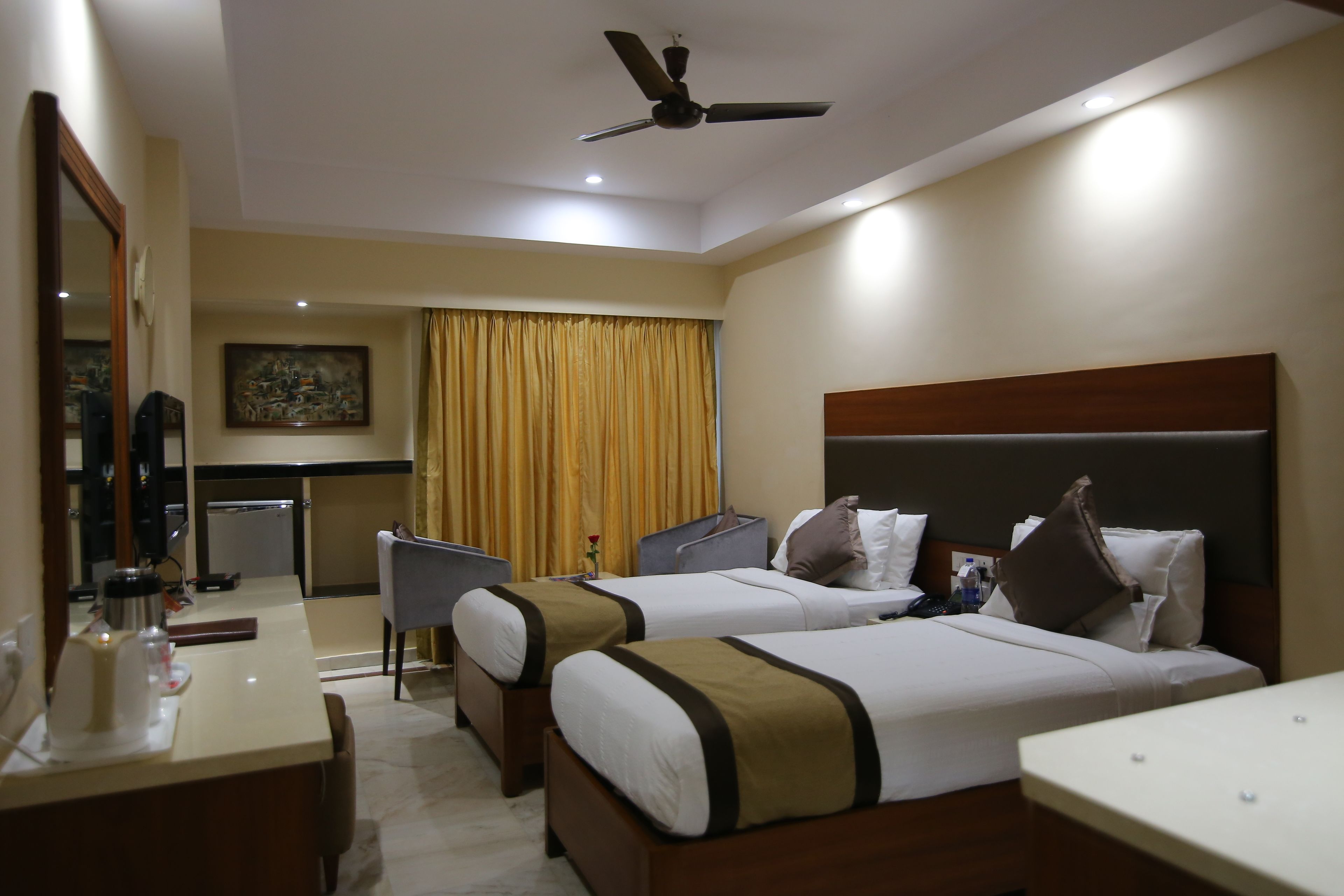 A hotel room with two beds, a desk, a chair, a ceiling fan, and a painting on the wall.