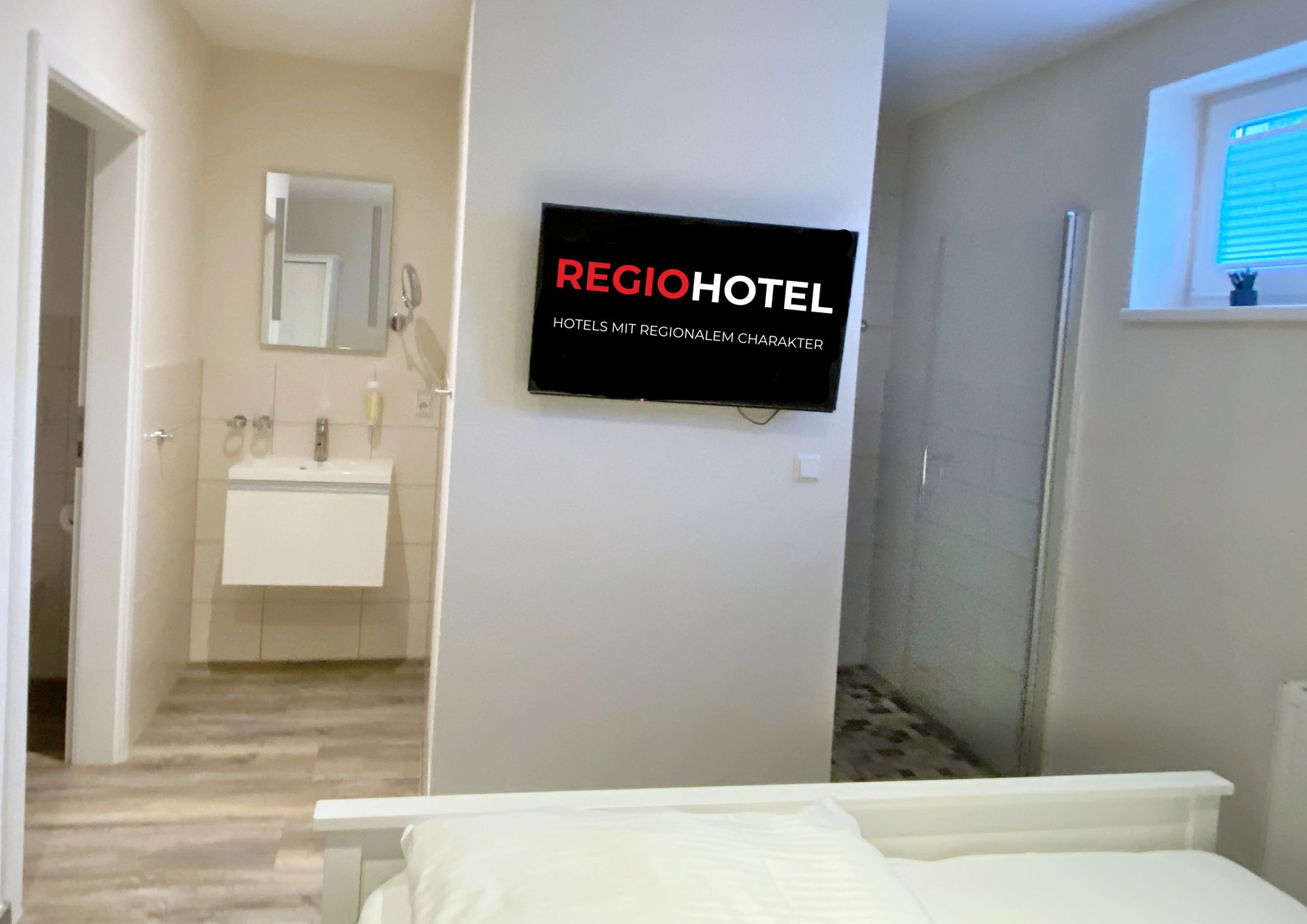 Ein Hotelzimmer mit Bett, Badezimmer mit Waschbecken und Spiegel sowie einem an der Wand montierten Fernseher, der 'REGIOHOTEL' anzeigt.