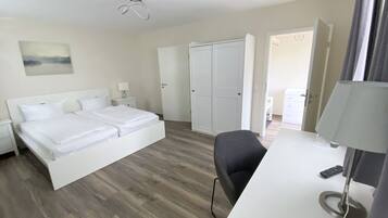 A hotel room with a bed, a desk, a chair, a desk lamp, a remote control, and a door leading to another room.