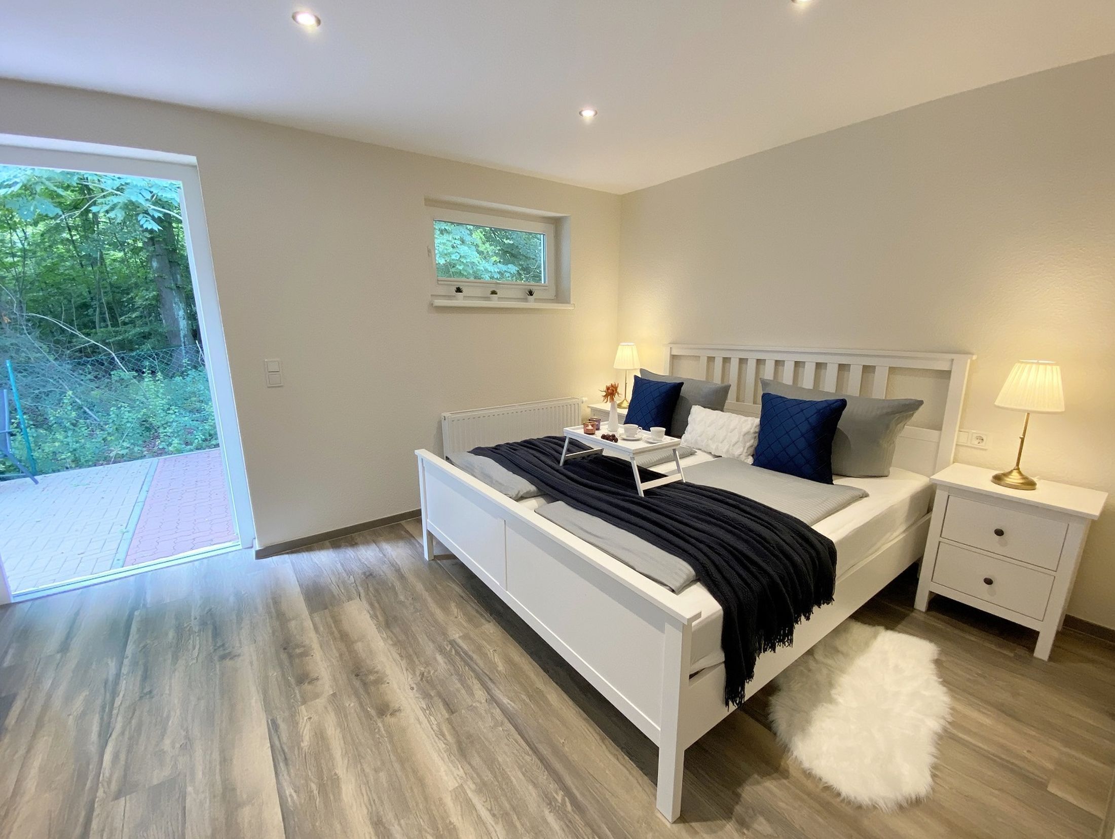 Une chambre moderne avec un lit blanc, deux tables de chevet, une petite fenêtre et une vue sur la verdure.